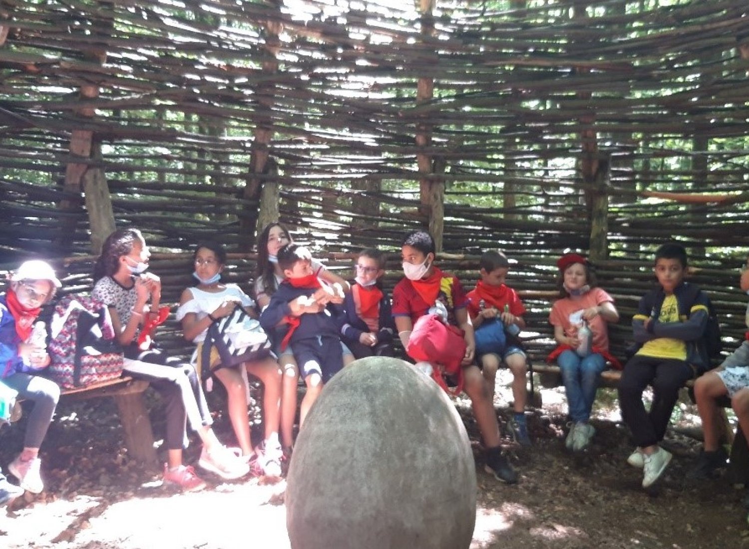Des enfants assis dans une hutte faite de branches autour d'un rocher  illuminés par la lumière du soleil passant à travers les branchages